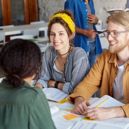 Back view of four students sitting together at table surrounded with books doing their common project sharing ideas with each other having pleasant smiles on their faces enjoying working together